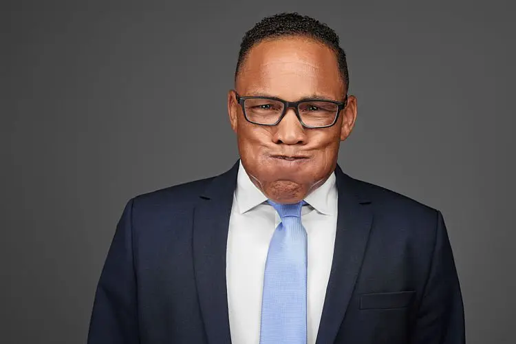 Creative team headshot of a man in glasses, a navy suit, and a light blue tie making a playful expression against a gray studio background.