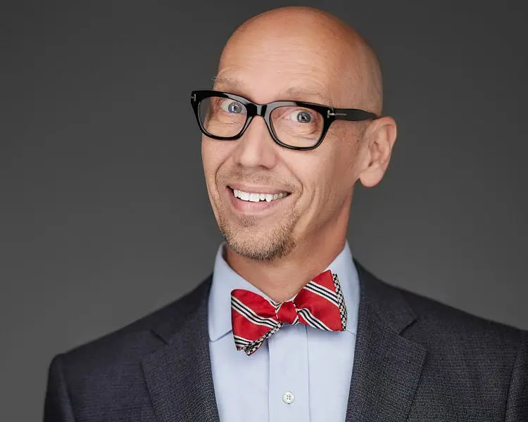 Professional team headshot of a smiling bald man wearing black glasses, a red bow tie, and a suit jacket against a gray background