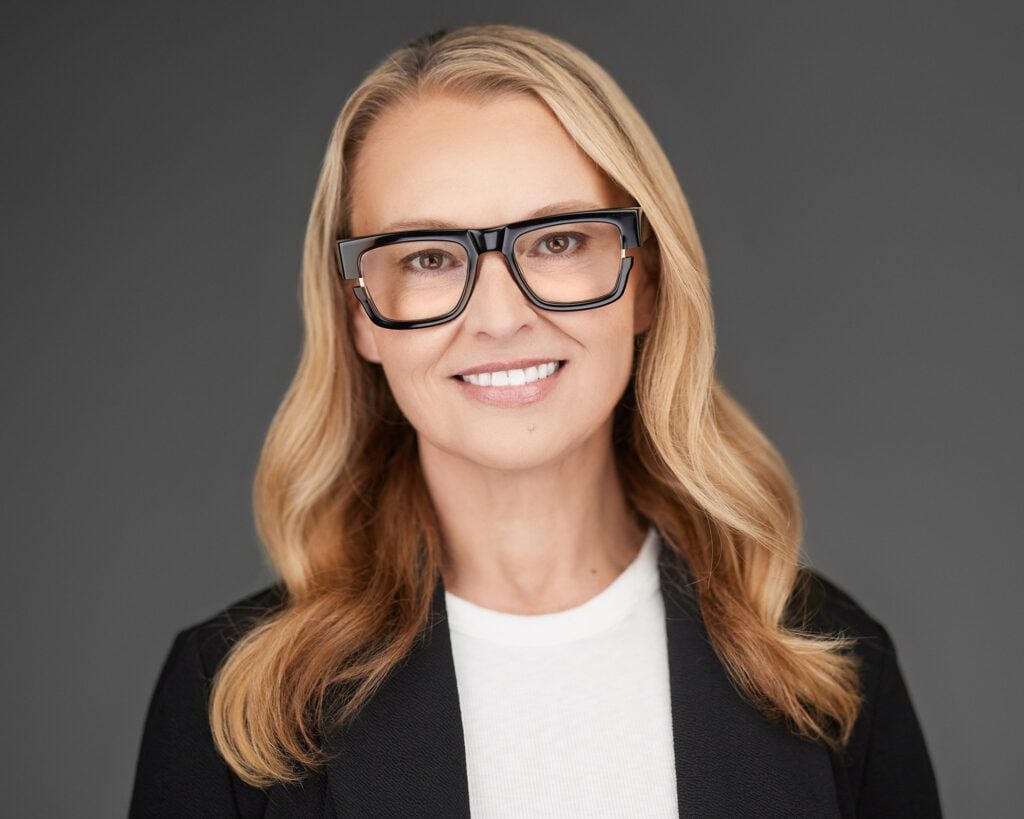 Professional team headshot of a blonde woman wearing bold black glasses, a black blazer, and a white top against a gray background