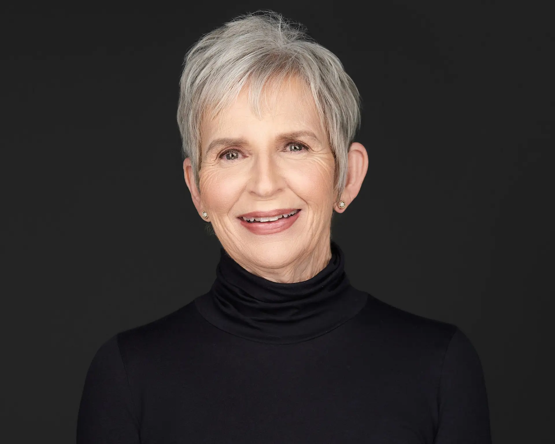 Executive team headshot of an judge woman with short gray hair wearing a black turtleneck against a dark studio background